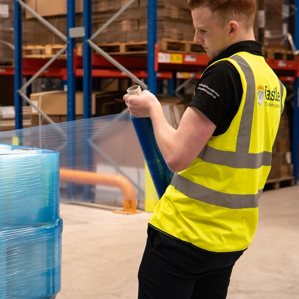 Warehouse worker wraps a pallet by hand in blue pallet wrap.