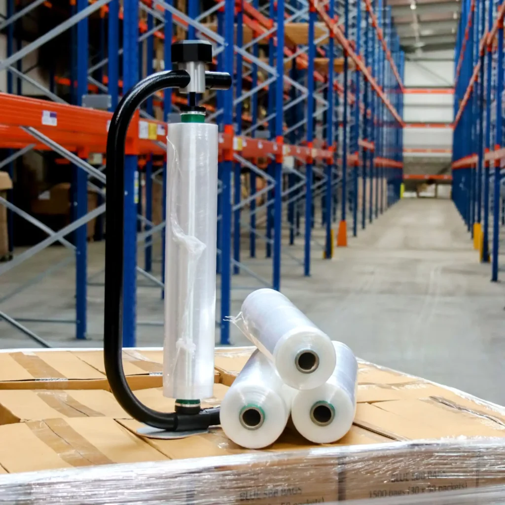 Stretch film roll attached to a hand film dispenser, with three film rolls laid next to it in a warehouse