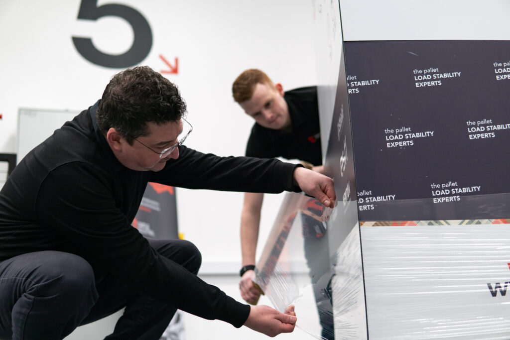 Paul and Tristan checking hand pallet wrap tension around a stacked pallet